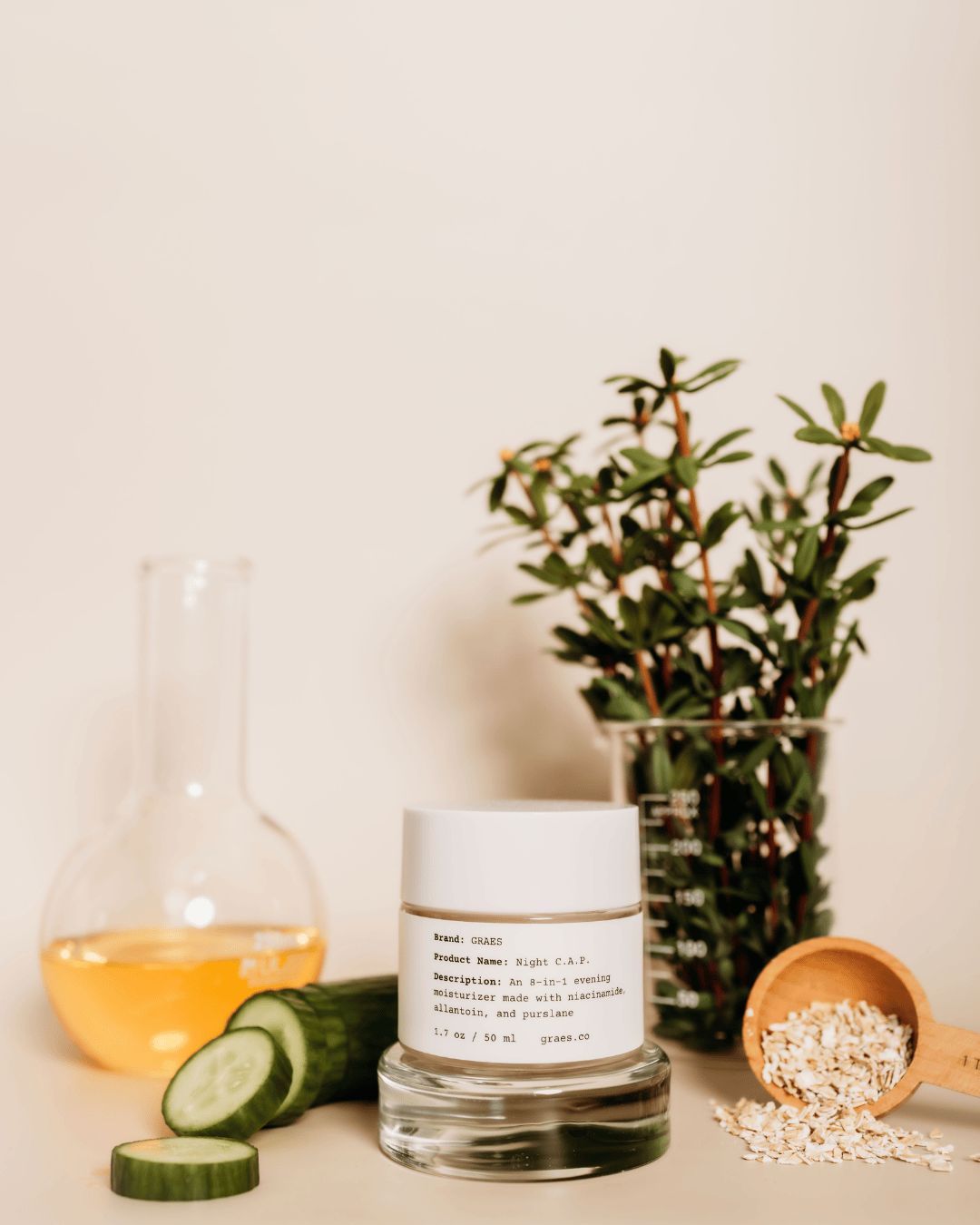 GRAES Night C.A.P. glass jar with label, sliced cucumber, wooden scoop with oatmeal, flask of jojoba oil, and purslane on a light surface.